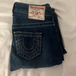 True Religion blå bootcut jeans - Snygga blå jeans från True Religion med klassisk kontrastsöm och ikonisk bakficka. Jeansen är tillverkade i mjukt denimtyg och har en tydlig logopatch baktill i rosa och vitt. De är endast använda fåtal gånger, med inga defekter! Pris går att diskutera, nypris: 1300kr 🙂