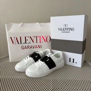 Valentino Garavani Rockstud sneakers - Säljer ett par snygga och HELT NYA Valentino Garavani Rockstud sneakers i vitt skinn med svart band och ikoniska pyramidnitar. Skorna har rund tå, platt sula med mönster och vita snören. Stl 37-38 Helt nya! Perfekt för dig som vill sticka ut med lyxig streetstyle. 100% äkta Andvända ca en vecka efter annonsen uppladdades
