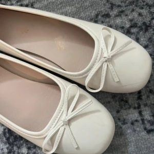 Vita ballerinaskor med rosett - Snygga vita ballerinaskor med rund tå och söt rosettdetalj framtill. Skorna har en klassisk och stilren design i syntetmaterial, perfekta för dig som gillar en clean och feminin look.