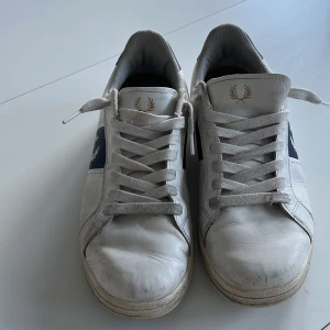 Fred Perry vita sneakers med blå detaljer - Snygga vita sneakers från Fred Perry med marinblå detaljer och logga på sidan samt hälen. Skorna har snörning, rund tå och platt sula. Tillverkade i skinn med klassisk siluett och stilren design som passar till jeans och hoodie.