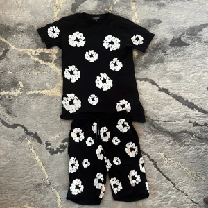 Svarta shorts med vita blommor - Säljer ett par svarta shorts med stora vita blommiga tryck. De har elastisk midja med snörning och är knälånga, perfekta för en chill och trendig look. Materialet känns mjukt och bekvämt.