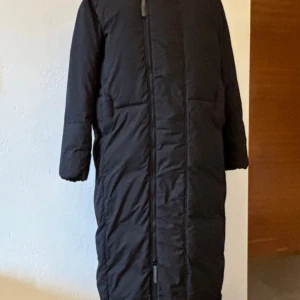 Svart lång kappa från G-Star RAW - Lång svart kappa från G-Star RAW med huva, tvåvägs dragkedja framtill, och diskret logga på höger och vänster sida på bröstet. Kappan har antingen en rak passform eller att man drar åt i midjan, slits med knappar nertill på båda sidor med knappar. Perfekt för kalla dagar när du vill hålla stilen och värmen. Stor i storleken.