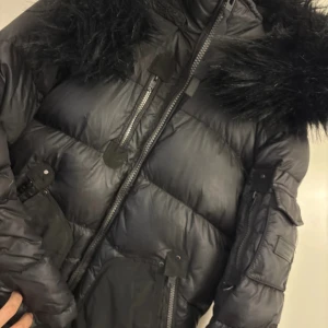 Svart dunjacka Rockandblue med fuskpäls - Säljer en svart dunjacka från Rockandblue med stor huva och svart fuskpäls. Jackan har flera fickor med dragkedja och tryckknappar, quiltad design och detaljer på ärmarna. Perfekt för kalla dagar och riktigt snygg streetstil.