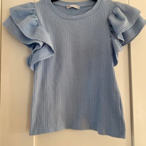 Blå ribbad topp med volangärm från Zara - Supersöt ljusblå topp från Zara med ribbad struktur och stora volangärmar. Toppen har rund halsringning och är tillverkad i ett mjukt, stretchigt material som känns skönt mot huden. Perfekt för dig som vill ha en trendig och lekfull look.  Mått Över bröstet: ca 35 cm Längd från axeln och ner: ca 49 cm  Polyester, bomull och elastan  Kommer från ett djur och rökfritt hem. 