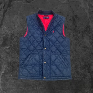 Quiltad blå väst från Polo Ralph Lauren - Quiltad väst från Polo Ralph Lauren i marinblått med röd insida. Västen har tryckknappar framtill, två fickor och en liten röd logga broderad på bröstet. Klassisk och sportig look med ribbad krage och snygg passform.
