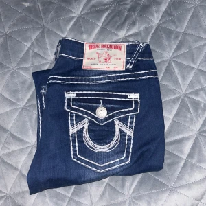 True Religion blå jeans med kontrastsömmar - Snygga blå jeans från True Religion med tydliga vita kontrastsömmar och ikoniska ficklock bak med silverknappar. Modellen har en rak passform och klassisk femficksdesign. Materialet är slitstarkt jeans och detaljerna gör dem riktigt unika.