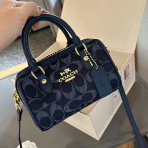 Coach handväska i olika färger - Säljer snygga handväskor från Coach i flera färger. Väskorna har klassisk boxig form, dubbla handtag, avtagbar axelrem och guldfärgade detaljer. Välj mellan marinblått, beige med bruna detaljer eller mörkbrun med svart. Coach-logga framtill och dragkedja upptill.