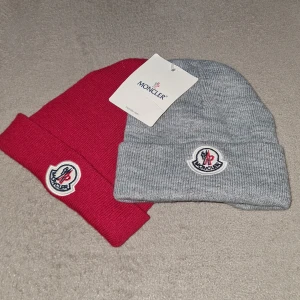 Moncler mössor i grått och rött - Två snygga Moncler mössor, en i grått och en i rött. Båda är ribbstickade med uppvikt kant och har Monclers ikoniska logga framtill. Perfekta för att hålla värmen och ge din outfit en exklusiv touch.