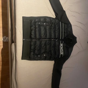 Svart pufferjacka från Moncler - Svart pufferjacka från Moncler med quiltad front, vita detaljer och broderad logga på bröstet. Jackan har långa ärmar i stickad kvalitet, dragkedja framtill och två fickor med tryckknappar. Perfekt för dig som vill ha en stilren och sportig look.