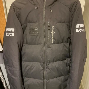 Svart dunjacka Sail Racing herr M - Svart dunjacka från Sail Racing med huva och reflekterande loggor på ärmar och bröst. Jackan har en quiltad design, dragkedja framtill och bröstficka med dragkedja. Perfekt för kalla dagar och har en clean, sportig look.