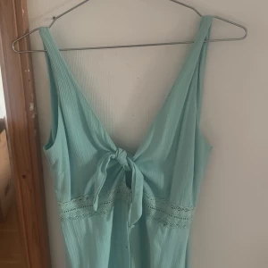 Ljusblå långklänning med knyt - Superfin ljusblå långklänning med knyt framtill och v-ringning både fram och bak. Klänningen har breda axelband, dekorativ spetsdetalj i midjan och knappar längs framsidan. Materialet är lätt och faller snyggt, perfekt för varma dagar.