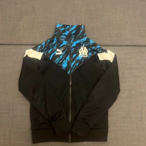 Puma Marseille zip-tröja - Säljer en svart och blå zip-tröja från Puma med Olympique de Marseille-logga broderad på bröstet. Tröjan har ett coolt mönster upptill, vita detaljer på axlarna och hel dragkedja. Perfekt för dig som gillar fotboll och vill ha en sportig look.