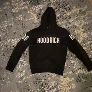Svart hoodie från Hoodrich med tryck - Svart hoodie från Hoodrich i storlek S med stor vit logga på bröstet och armarna. Tröjan har huva med snörning, långa ärmar och en ficka med dragkedja på sidan. Materialet är mjukt och känns som bomull och polyester. Perfekt för dig som gillar streetwear.