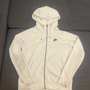 Beach hoodie från Nike med dragkedja - Säljer en stilren beach hoodie från Nike med svart logga på bröstet. Tröjan har huva med snörning, praktiska fickor framtill och hel dragkedja. Materialet är mjuk bomull och polyester, perfekt för chill eller träning. Långärmad och normal passform.