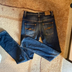 Mörkblå  jeans från Nudie - Säljer ett par mörkblå jeans från Nudie med klassiska kontrastsömmar och snygga detaljer på bakfickorna. Jeansen har fem fickor, normal midja och är tillverkade i stretchig bomullsdenim för en bekväm passform.