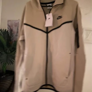 Beige Nike vindjacka med svarta detaljer - Nike vindjacka i beige med svarta linjer och dragkedja framtill. Jackan har hög krage, två sidofickor och Nike-logga på bröstet. Snygg sportig look med stilrena kontraster, perfekt för dig som gillar streetwear och sport.