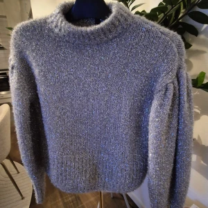 Glittrig grå stickad tröja - Supermysig grå stickad tröja med glittriga inslag och ribbade muddar. Tröjan har en klassisk rund halsringning och långa ärmar med lätt puff vid axlarna. Perfekt för dig som vill sticka ut lite extra med en glittrig touch.