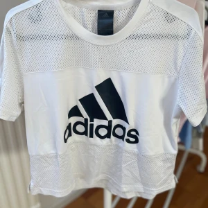 Vit mesh t-shirt från Adidas - Vit t-shirt från Adidas med svart logga framtill. T-shirten har korta ärmar och är tillverkad i ett lätt meshmaterial upptill och nedtill för extra ventilation. 