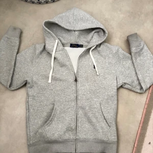 Grå hoodie från Polo Ralph Lauren - Grå hoodie från Polo Ralph Lauren med dragkedja, vita snören och klassisk broderad logga på bröstet. Mjuk bomullsmix, kängurufickor och ribbade muddar. Perfekt för chill dagar eller när du vill ha en clean och enkel look.