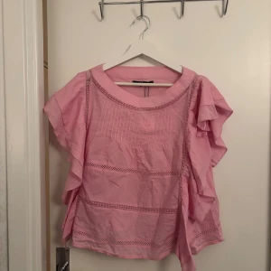 Rosa blus med volangdetaljer - Säljer en söt rosa blus med volangdetaljer på ärmarna och dekorativa spetsband framtill. Blusen har rund halsringning och är tillverkad i mjuk bomull, perfekt för en chill och feminin look. Passar dig som gillar romantisk stil och ljusa färger.