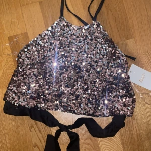 Onesize glittrig paljett topp med svarta sidenband - Säljer en glittrig topp med paljetter. Toppen knyts i nacke och rygg, så storleken kan justeras. Passar XS, S, M, L beroende på hur man knyter det. Perfekt till fest, nyår eller en utekväll. Helt nytt, aldrig använt och med tag kvar. 
