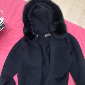 Alyssa jacka - Svart stickad cardigan jacka från Alyssa med fluffig svart fuskpäls runt huvan. Jackan har långa ärmar, ribbade muddar och dragkedja framtill. Perfekt för dig som gillar mysiga och stilrena plagg med extra detaljer. Its basically new only worn 2times