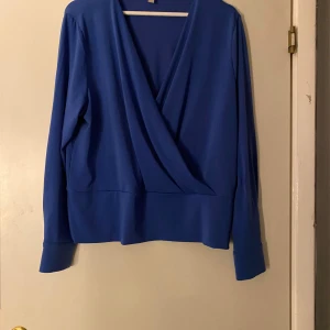 Blå omlottblus med guldfärgade knappar - Snygg blå omlottblus från H&M med lång ärm och breda manschetter med guldfärgade knappar. Blusen har en djup v-ringning och ett brett band nedtill som ger en cool siluett. Perfekt att styla med jeans eller kjol för en trendig look.