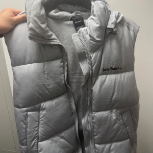 Ljusgrå dunväst från iets Frans - Snygg ljusgrå dunväst från iets Frans med huva och dragkedja framtill. Västen är quiltad och har två fickor på sidorna samt en hög krage. Perfekt för lager-på-lager och streetstyle.