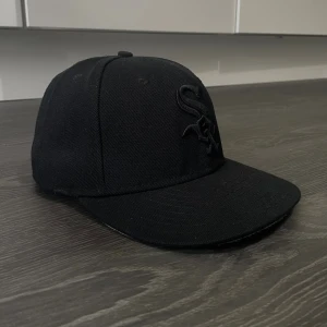 Svart New Era 59FIFTY White Sox keps - Säljer en svart New Era 59FIFTY keps med broderad White Sox-logga framtill. Klassisk flat peak och broderade detaljer i svart. Kepsen är i bomull och polyester, med MLB-logga bak och New Era-logga på sidan. Perfekt för dig som gillar streetwear och sport.