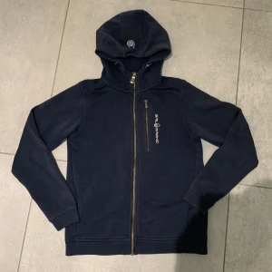 Marinblå hoodie från Sail Racing - Säljer en marinblå hoodie från Sail Racing med hel dragkedja och huva. Tröjan har en snygg logga på bröstet och en praktisk bröstficka med dragkedja. Perfekt för dig som gillar sportig och stilren look.