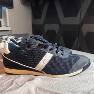 Marinblå sneakers från Hugo Boss - Snygga marinblå sneakers från Hugo Boss med vit detalj på hälen och sidan. Ovandelen är i mesh och mocka, med svarta snören och vit sula. Klassisk och stilren design med BOSS-logga på sidan och hälen. Perfekta för dig som gillar en sportig men clean look.