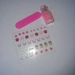 Nageldekorationer med rosa glitter - Sött kit med nageldekorationer: ark med klistermärken i form av kaniner, tassar, blommor och stjärnor i rosa, gult och svart. Inkluderar en liten glasflaska med rosa glitter och en rosa minifil. Perfekt för att göra dina naglar extra gulliga och kreativa.