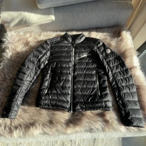 Svart Moncler dunjacka Longue Saison - Svart dunjacka från Moncler, modell Longue Saison. Jackan har quiltad design, ståkrage och dragkedja framtill samt tre fickor med dragkedja. Klassisk Moncler-logga på ärmen. Perfekt för kyliga dagar och riktigt snygg passform. Pris 3000kr mennksn diskuteras.