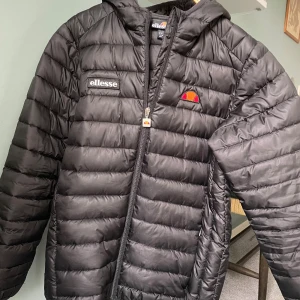 Svart pufferjacka från Ellesse - Svart pufferjacka från Ellesse med huva och dragkedja framtill. Jackan har Ellesse-logga på bröstet och är quiltad med horisontella sömmar. Perfekt för kyliga dagar och har en sportig vibe.