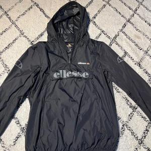 Svart vindjacka från Ellesse - Svart vindjacka från Ellesse med stor logga på bröstet och mindre logga på ärmen. Jackan har huva, lång ärm och en praktisk ficka framtill. Materialet är lätt och i syntet, perfekt för blåsiga dagar. Halv dragkedja framtill för enkel påtagning.