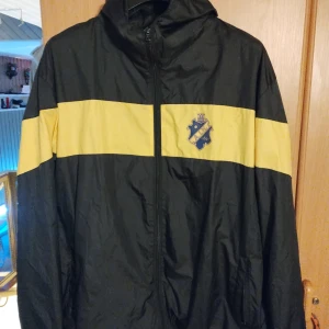 AIK vindjacka svart/gul 2XL - Snygg svart vindjacka från AIK med gul horisontell rand över bröst och armar. Jackan har huva, dragkedja framtill och AIK:s klubbmärke på bröstet. Perfekt för dig som vill visa ditt stöd för laget och hålla stilen på läktaren.