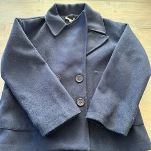 Mörkblå skepparkavaj från Design by S - Stilren mörkblå skepparkavaj från Design by S i storlek XS/S. Jackan har dubbelknäppning med stora knappar, klassisk krage och raka ärmar. Materialet är mjukt och känns ulligt, perfekt för kyligare dagar. Enkel och tidlös design som funkar till många outfits. Lite nopprig på ärmarna. Kan säkert fixas. 