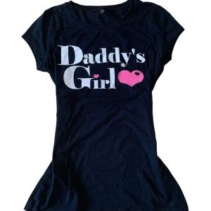 Vintage Svart baby tee Daddy's Girl Fishbone - Svart t-shirt från Fishbone med vit och rosa text 'Daddy's Girl' och hjärtdetalj framtill. Klassisk rund halsringning och korta ärmar. Skön passform i mjuk bomull, perfekt för en chill och lekfull stil.