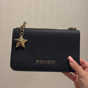 Marinblå plånbok från Tommy med stjärndetalj - Snygg marinblå plånbok från Tommy med guldfärgad stjärnberlock och logga framtill. Plånboken har en stilren, rektangulär form och insida i ljus färg. Perfekt för dig som vill ha något både trendigt och praktiskt.