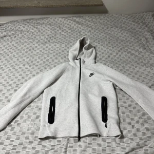 Ljusgrå nike tech - Nike hoodie i ljusgrått med svart logga på bröstet. Tröjan har hel dragkedja framtill, två svarta dragkedjeförsedda fickor och huva. Mjuk och skön, perfekt för chill eller träning. Långärmad och i klassisk normal passform.