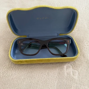 Bruna glasögon från Gucci - Snygga glasögon från Gucci med rektangulära bågar i mörkbrunt acetat. Guldiga GG-detaljer på skalmarna ger en lyxig touch. Kommer i originalfodral med blå insida och gul sammet på utsidan. Perfekt för dig som vill ha en stilren och exklusiv look.