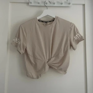 Beige croppad t-shirt från Stronger - Säljer en beige croppad t-shirt från Stronger i storlek XS. Toppen har knutdetalj framtill, korta ärmar och tryckt logga på båda armarna. Tillverkad i mjuk och stretchig polyesterblandning, perfekt för träning.