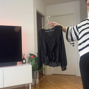 Svart blus med volang och knappar - Säljer en svart blus med v-ringning, volangdetaljer framtill och knappar längs hela framsidan. Blusen har långa ärmar med breda muddar och är tillverkad i ett lätt, flowy material som ger en snygg siluett.