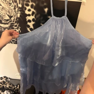 Ljusblå volangklänning med knytband & öppen rygg  - Supersöt ljusblå klänning med flera lager volanger och tunna knytband i nacken. Klänningen är ärmlös och har en glansig, lätt transparent look som ger en drömmig känsla. Perfekt för dig som vill sticka ut med en romantisk och luftig stil.