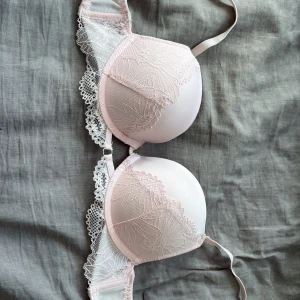 Ljusrosa spets-bh med blommönster - Supersöt ljusrosa bh med vadderade kupor och vacker spets med blommönster. Axelbanden är justerbara och ryggen har klassisk hysk- och hakknäppning. Perfekt för dig som gillar romantiska detaljer och mjuka pasteller.