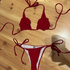 Röd bikini med rosettdetaljer - Snygg röd bikini med knyt i både över- och underdel. Överdelen har dekorativa blommor i tyg på kuporna som ger en unik och trendig look. Perfekt för stranden eller poolen i sommar! Använts 1 gång. Passar XS/S