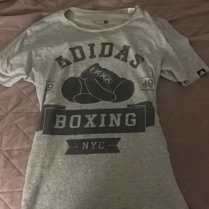 Grå Adidas Boxing t-shirt - Snygg grå t-shirt från Adidas med stort svart tryck framtill med boxningshandskar och texten 'Boxing NYC'. Klassisk rund halsringning och korta ärmar. Perfekt för dig som gillar sportig streetstyle.