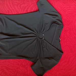 Svart topp med ringdetalj från BikBok - Svart kortärmad topp från BikBok med snygg ringdetalj och rynk mitt fram. Toppens material känns mjukt och stretchigt, perfekt för en trendig look. Passformen är normal och den har en enkel, stilren design som passar till mycket.