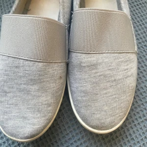  Gråa skor och   - Snygga gråa slip-on loafers med elastiskt band över foten. Skorna har en platt vit sula och är tillverkade i mjuk bomullstextil, vilket gör dem riktigt bekväma och lätta att bära. Perfekta för dig som gillar en enkel och stilren look.