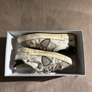 Philippe Model vita sneakers i skinn - Snygga vita sneakers från Philippe Model Paris med klassisk låg profil. Skorna är i skinn med svarta detaljer på häl och sula, samt märkets ikoniska sköld på sidan. De har snörning och en platt sula, perfekt för en avslappnad och trendig look.